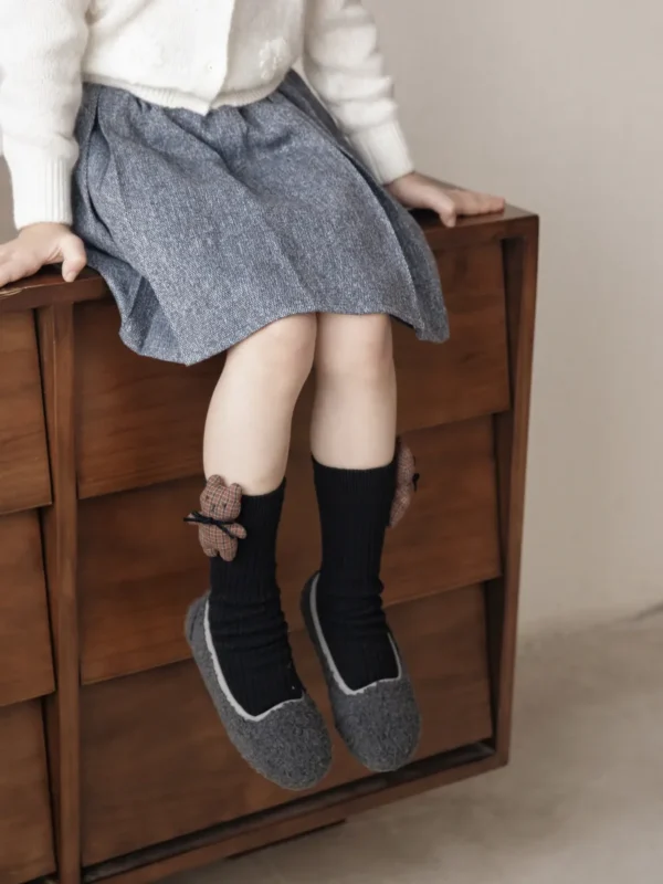 Calcetines negros con detalle de oso y lazo, combinados con una falda gris y unas acogedoras zapatillas grises.
