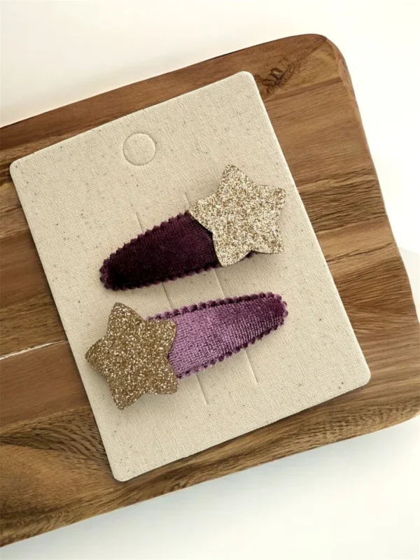 Two velvet hair clips in shades of purple with glittery gold star embellishments, displayed on a fabric backing.