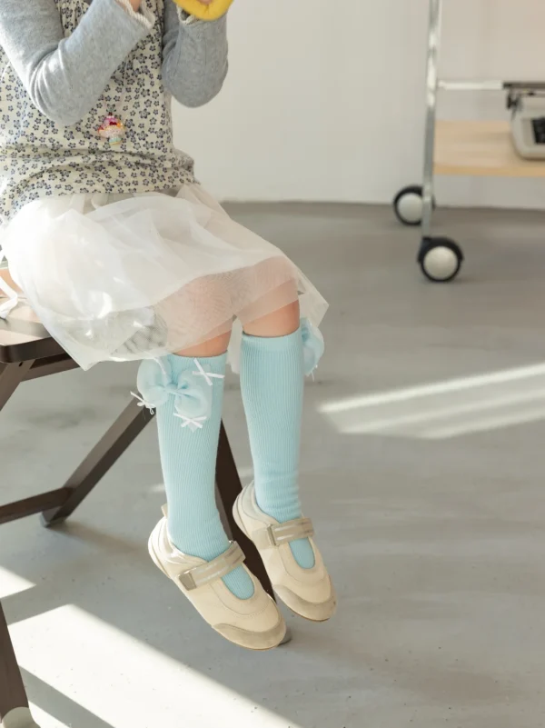 Niña con falda blanca y calcetines azules hasta la rodilla con lazos, con zapatos beige.