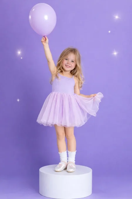 Niña con vestido morado sosteniendo un globo, sonriente, sobre fondo lila claro, con bailarinas y calcetines.